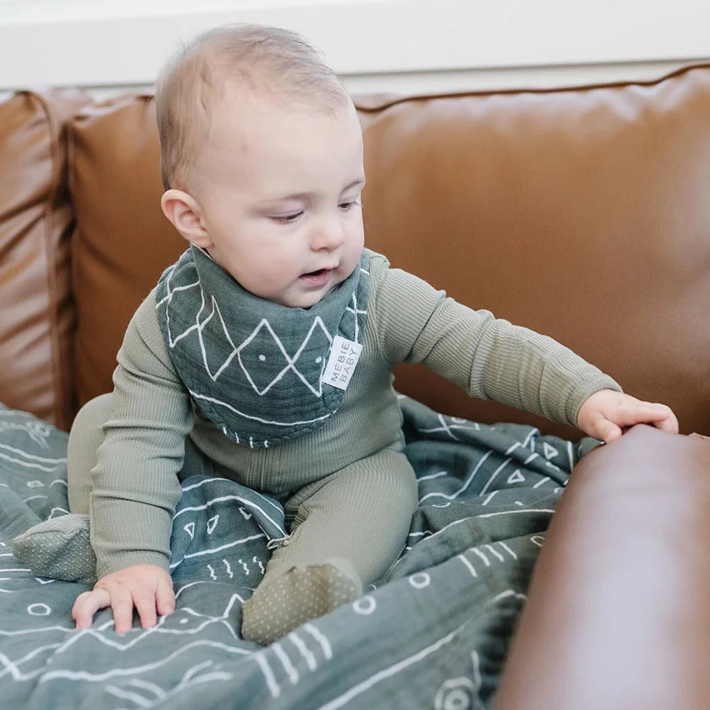 Baby sitting on leather couch, wearing Alpine Bib by Mebie Baby. Bib is a triangle bib with a white fabric tag that says "Mebie Baby", bib is a deep green colour with an aztec pattern.