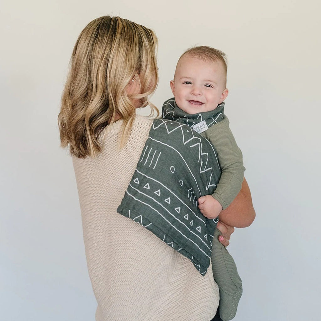 White background with mama holding baby, her back is to the camera, with an Alpine Burp Cloth by Mebie Baby over her shoulder. Burp cloth is made of muslin, is rectangular, green with an aztec print, and has a white tag that says "MEBIE BABY".