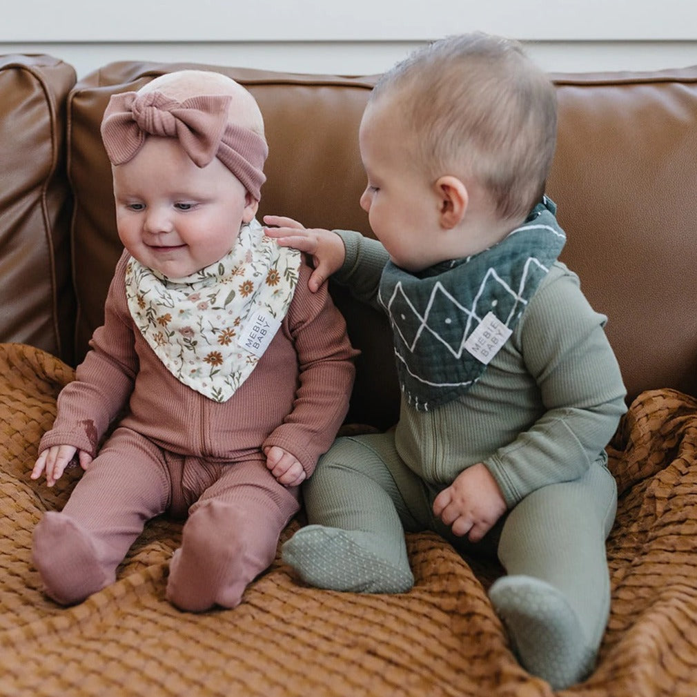 two babies sitting on a leather couch together, one of them is wearing the Meadow Floral Bib by Mebie Baby.
