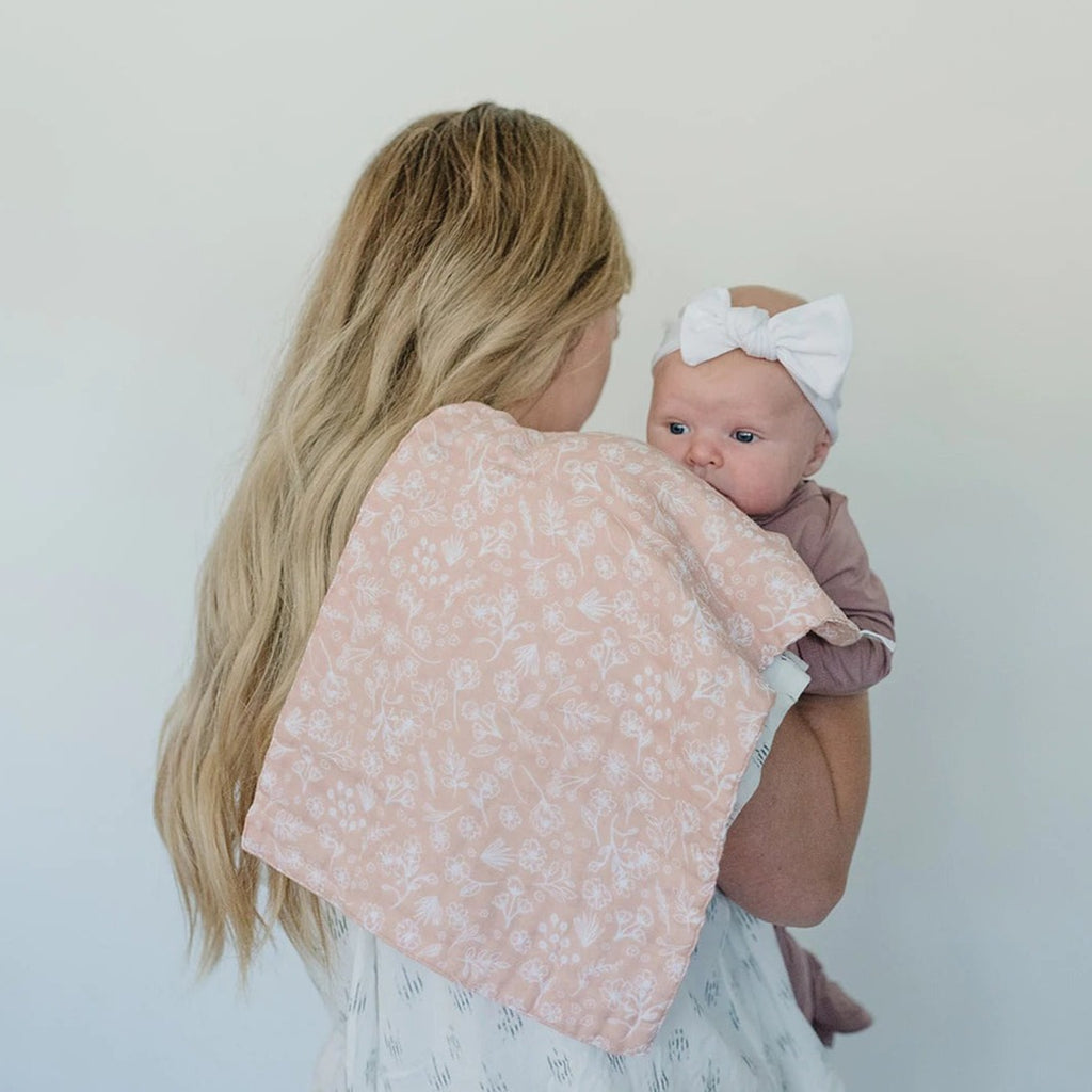 White background with a mama holding her baby, back to camera, with a Wildflower Burp Cloth by Mebie Baby draped over her shoulder. Burp cloth is a soft pink colour with white wildflowers all over.