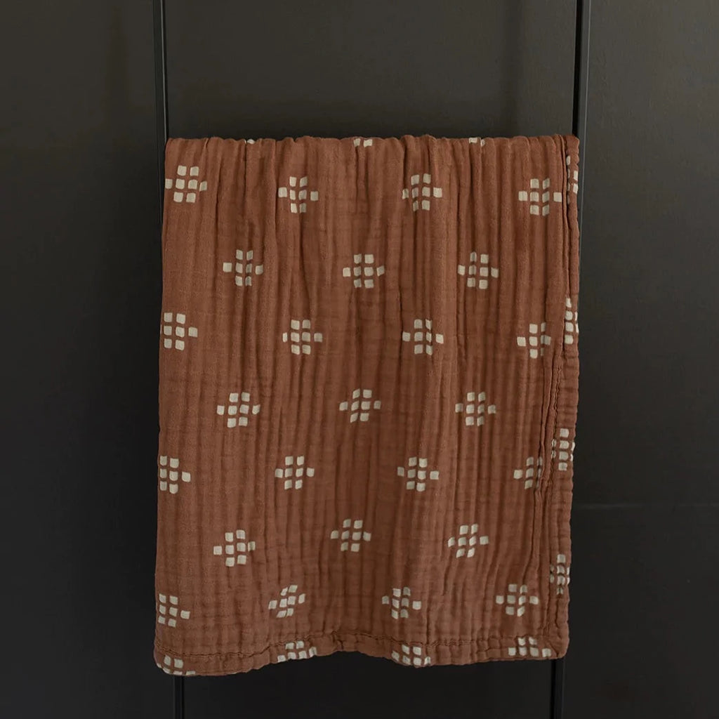 Dark background with a black metal blanket ladder with a Chestnut Textile Muslin Quilt by Mebie Baby. Quilt is dark brown with white/cream square pattern all over.