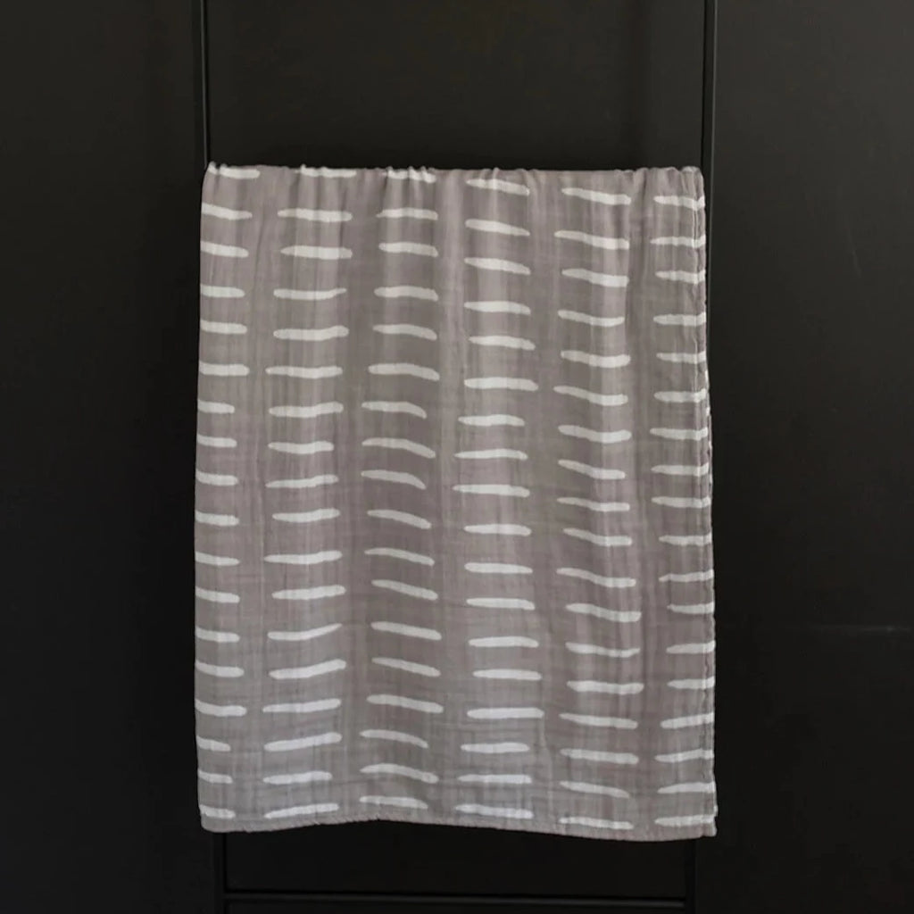 Dark background with a m=black metal blanket ladder, and a Grey Dash Muslin Swaddle by Mebie Baby hanging. Swaddle is a medium grey with white dashes all over.