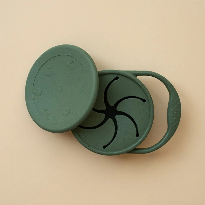Beige background with a Silicone Snack Cup in Leaf by Minika. Snack cup has a small handle to carry, and a lid as well, all in the colour emerald green speckled.\