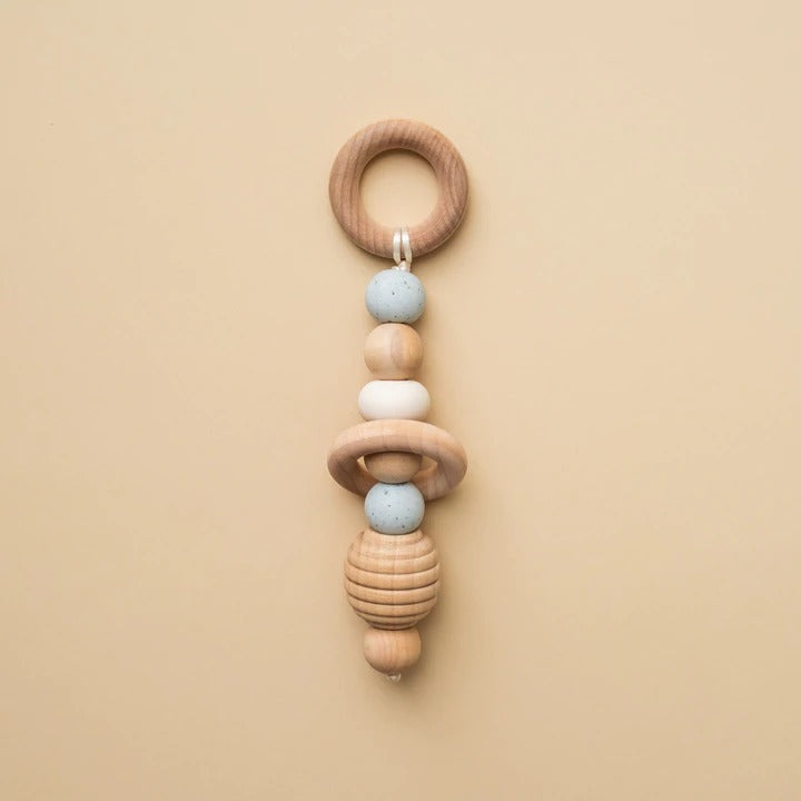 Beige background with a Hand Rattle in Ice by Minika. Hand rattle has a wooden circle with ice blue speckled silicone beads, white silicone and wood beads going down, and a honeycomb bead.