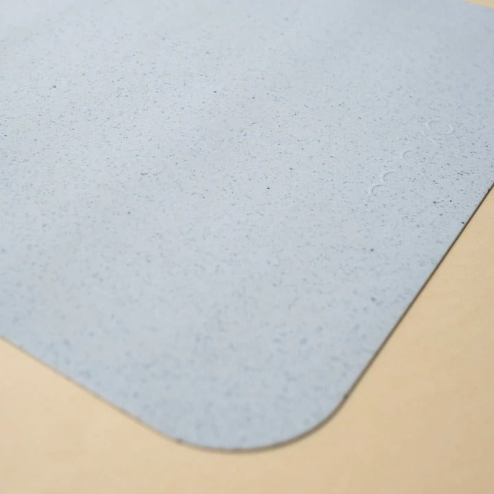 Beige background with a Silicone Placemat in Ice by Minika. Placemat is square silicone, in an ice blue speckled colour.