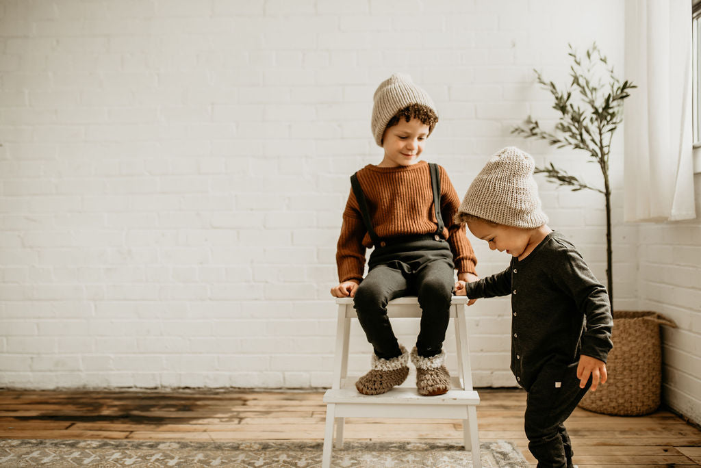 White brick wall with 2 boys standing, wearing the Handknint Beanie by Petit Nordique in Oat.