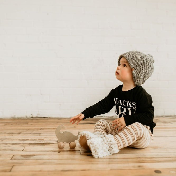 White brick wall with a little boy sitting on the ground, wearing the Handknit Teddy Booties by Petit Nordique.
