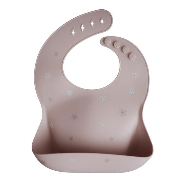 White background with a Silicone Bib in Blush Daisy by Mushie. Bib is a blush colour with white daisies, with a deep pocket on the front, and a rounded neck fastener at the back.