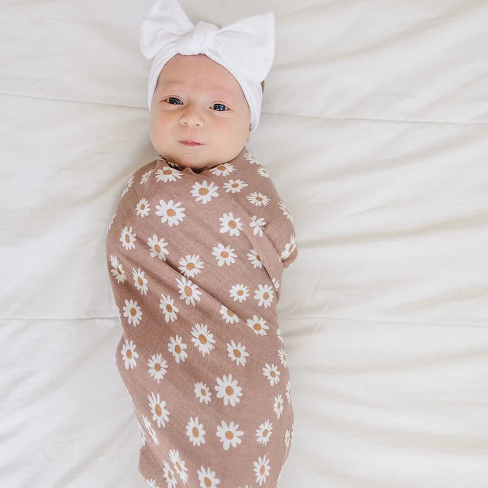 White background with a baby girl wearing a large white knot bow, and swaddled in a Daisy Dream Muslin Swaddle by Mebie Baby.