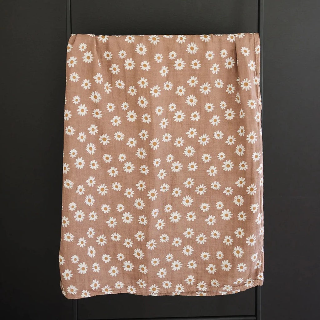 Dark background with a metal blanket ladder, and a Daisy Dream Muslin Swaddle by Mebie Baby hanging over it. Swaddle is a mauve colour with white daisies all over.