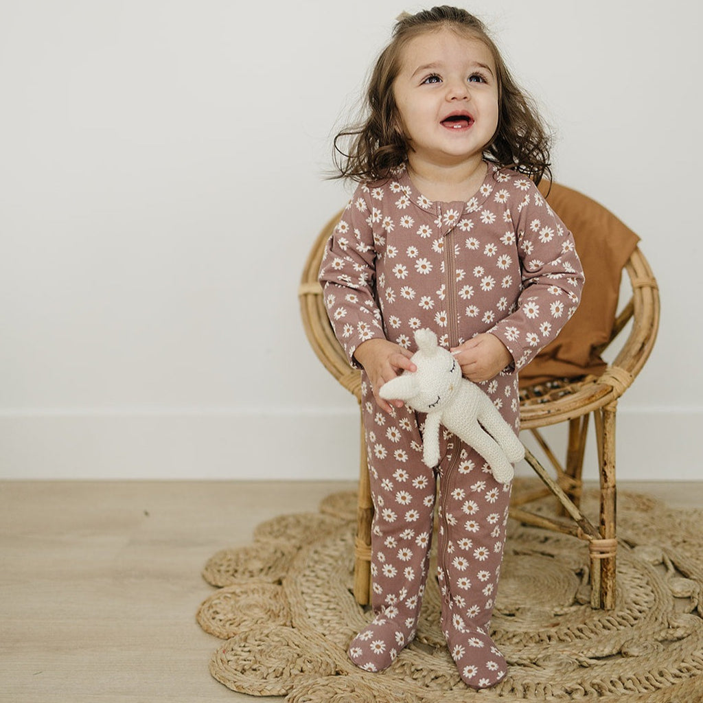 Daisy Dream Zipper by Mebie Baby on a little girl, standing in front of a rattan chair and a wicker carpet. 