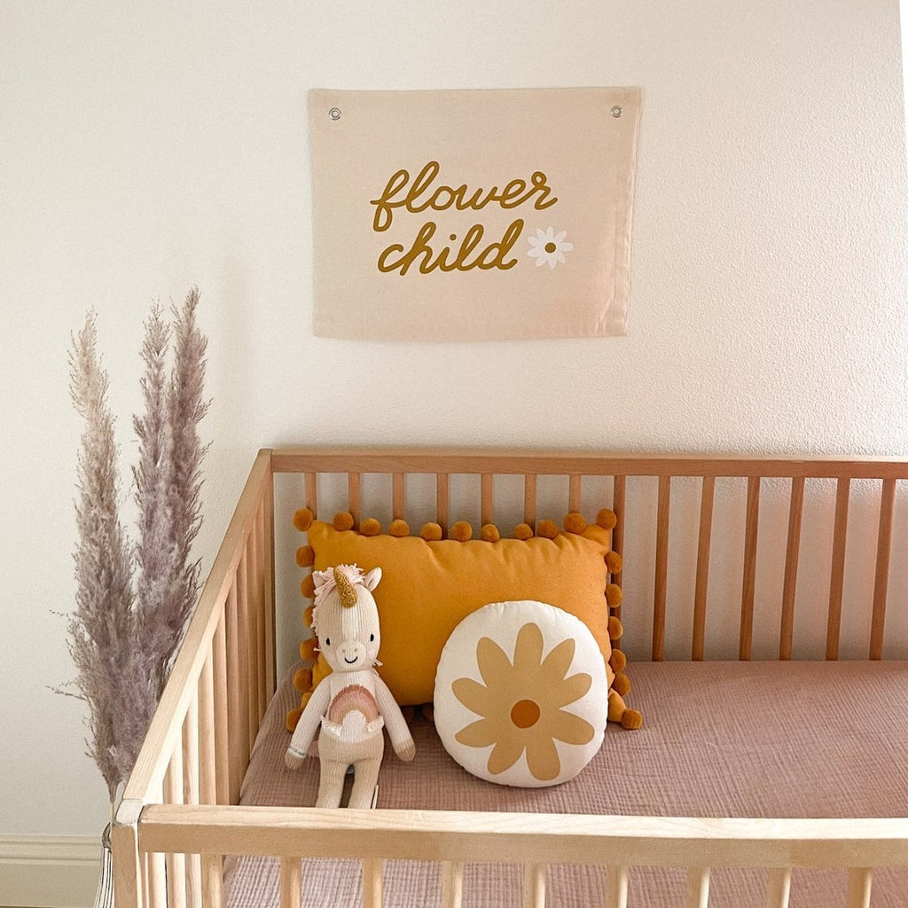 Overhead shot of a natural wood crib, with a banner on the wall, a unicorn stuffed animal and the Daisy Pillow by Imani Collective. Daisy pillow is a cream linen with a drawn on yellow daisy.