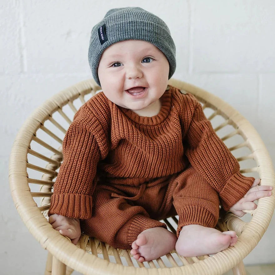 White brick wall with a rattan chair, and a baby sitting in it wearing the Dark Grey Beanie by Mebie Baby.