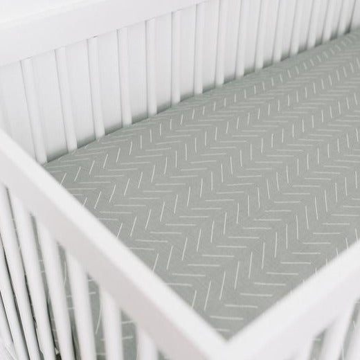 Side angle of white crib with Desert Sage Crib Sheet by Mebie Baby. Crib sheet is a pale sage colour with white lines all over, and fits snug to the mattress.