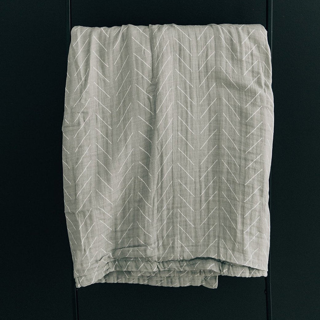 Dark background with a black metal blanket ladder, and a Desert Sage Muslin Quilt by Mebie Baby hanging over it. Quilt is a pale sage colour, with white lines.