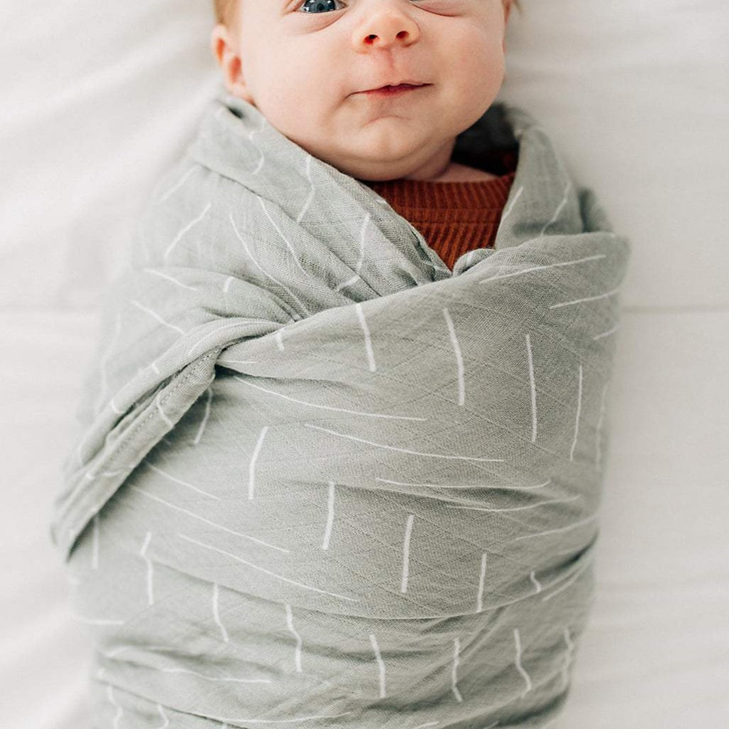 White background with a close up of a baby wrapped in a Desert Sage Muslin Swaddle by Mebie Baby. Swaddle is a pale sage colour, with white lines all over.