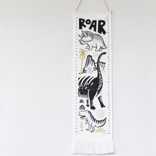 White background with the Dino Growth Chart by Wee Gallery hanging. Growth chart is white, and rectangular, with dinosaurs going down and says "ROAR" on the top, all in black, with yellowish accents.