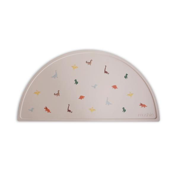 White background with Silicone Place Mat in Dinosaurs by Mushie. Placemat is beige with colourful dinosaurs, made of silicone, and in a semicircle.