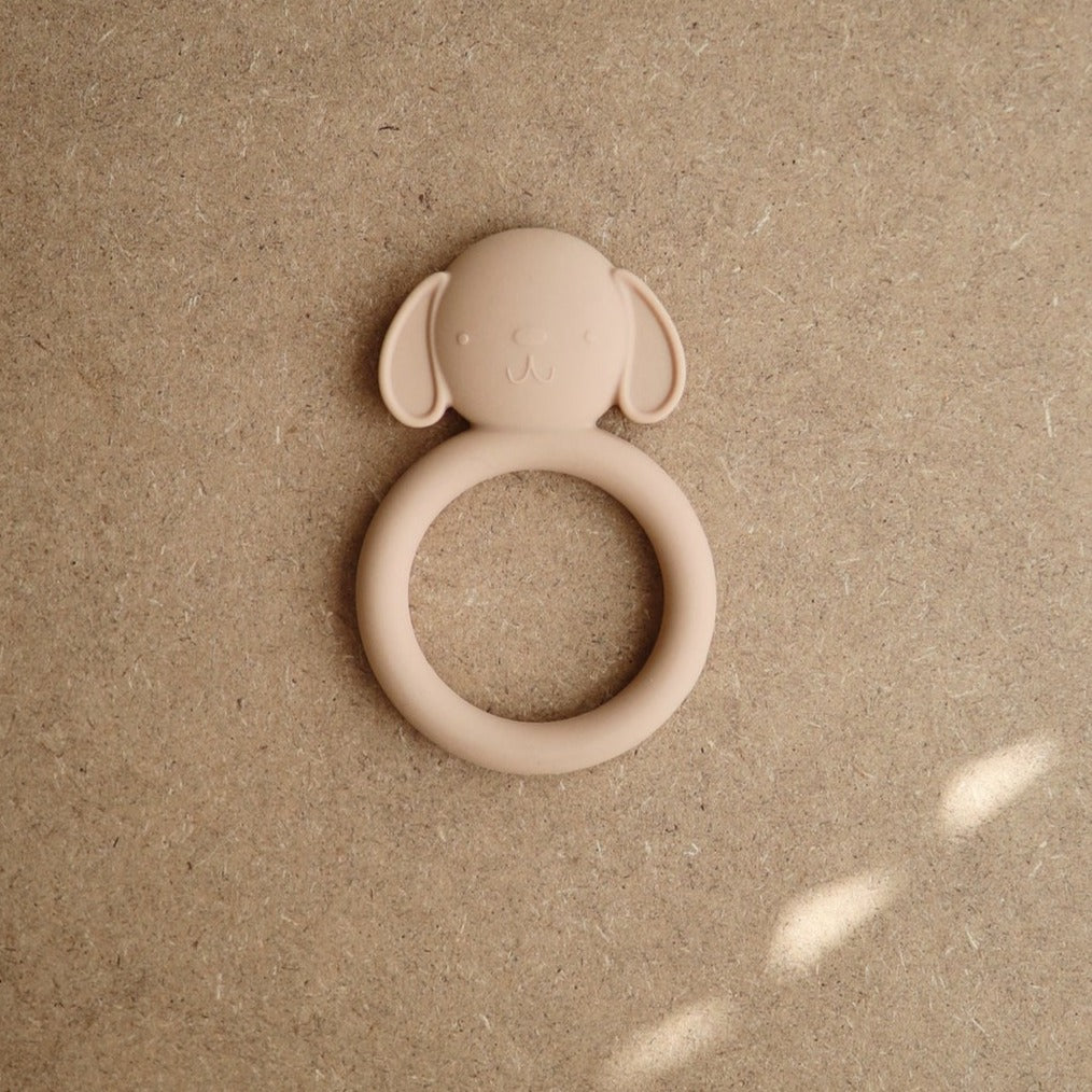 Brown background with the Dog teether by Mushie. Dog teether is beige with a round handle and a dog head, with floppy ears, all made out of food grade silicone.