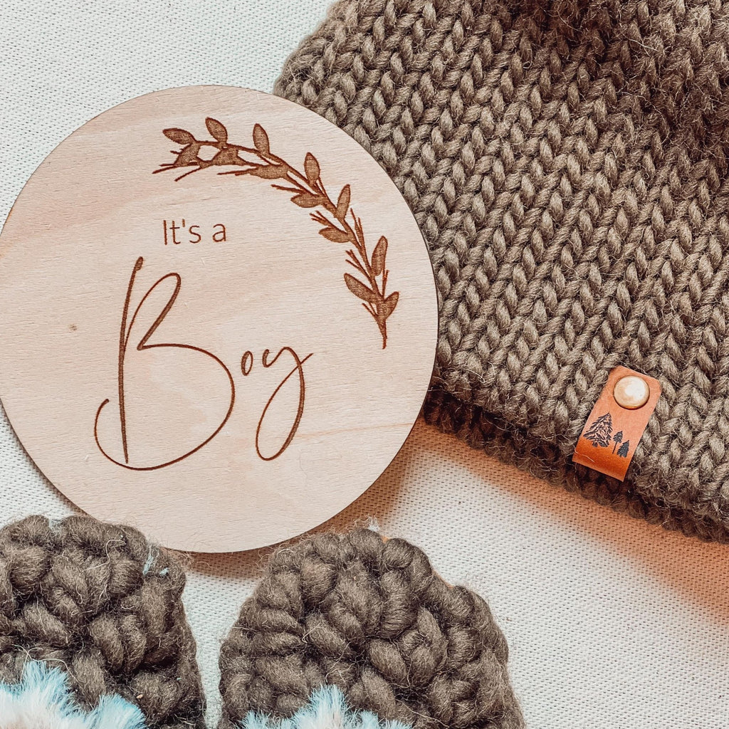 It's a Boy Double-Sided sex announcement milestone disc by Petit Nordique paired with a brown tweed beanie and taupe moccs by Petit Nordique, laid on a beige surface. 