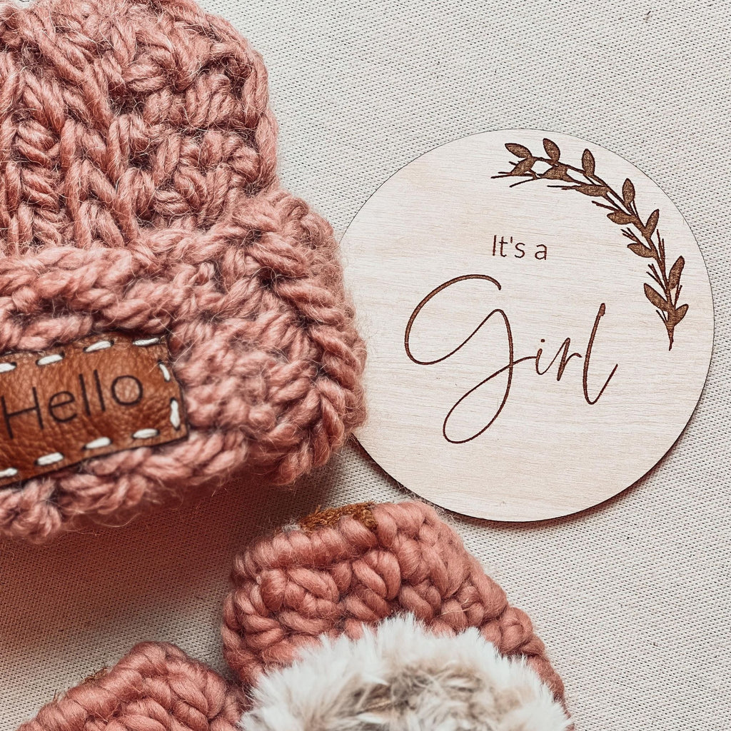It's a Girl Double-Sided sex announcement milestone disc by Petit Nordique paired with a Blush Take Me Home Beanie and Dusty Rose Moccs by Petit Nordique. Laid on a beige surface. 