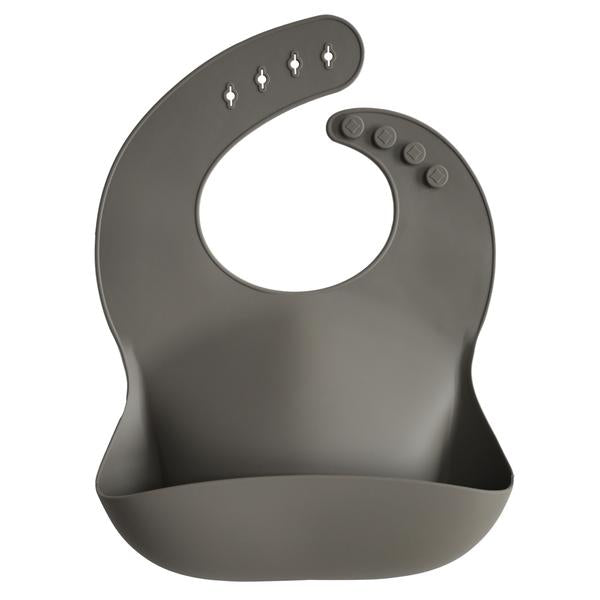 White background with a Silicone Bib in Dove Gray by Mushie. BIb is a dark gray colour with a deep pocket on the front, and a rounded neck fastener at the back.