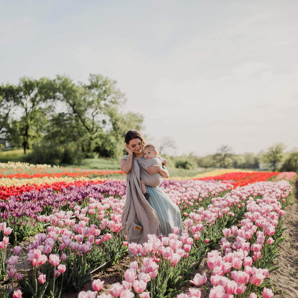 Woman standing outside, in a field of tulips, looking down at baby in a Ring Sling in Driftwood by Kyte Baby. Ring Sling is a neutral grey colour.