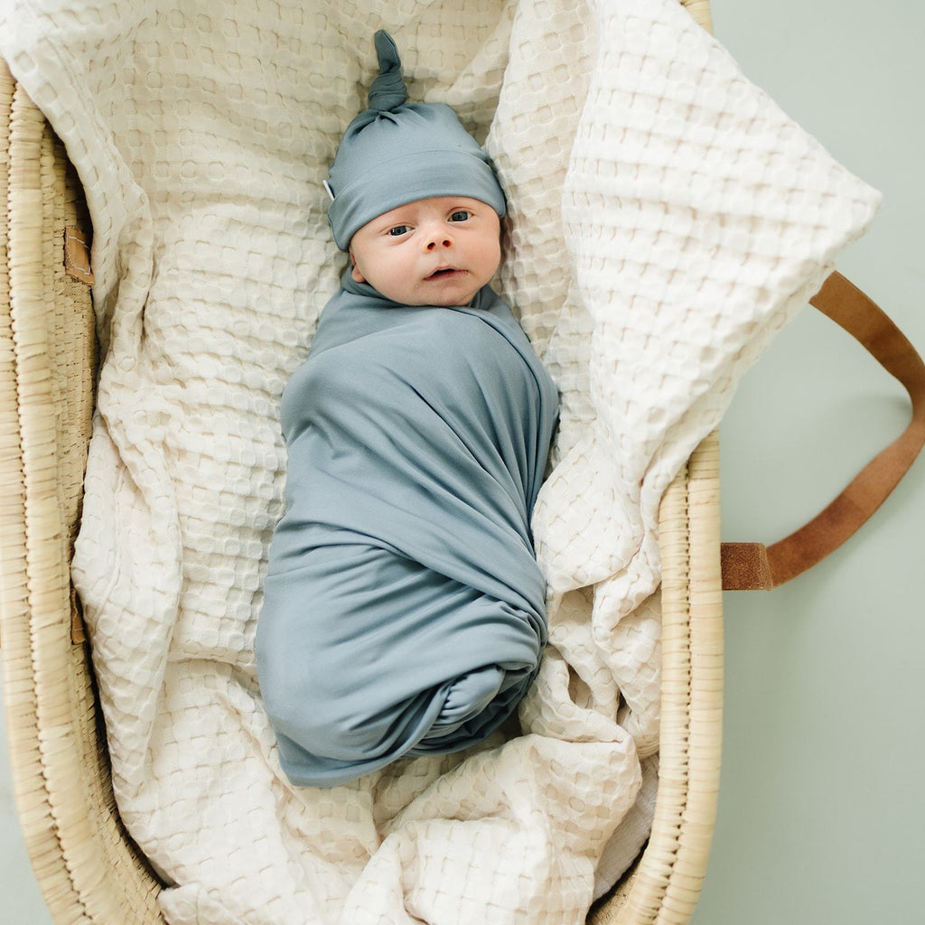 Overhead view of baby laying in a bassinet with a blanket, wrapped up in a Dusty Blue Stretch Swaddle by Mebie Baby. Swaddle is dusty blue, and full of stretch.