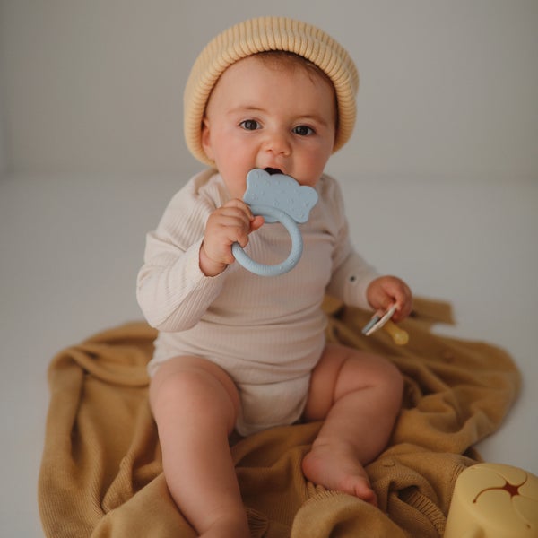 White background with a baby sitting on an ochre blanket, chewing on an Elephant Teether by Mushie.