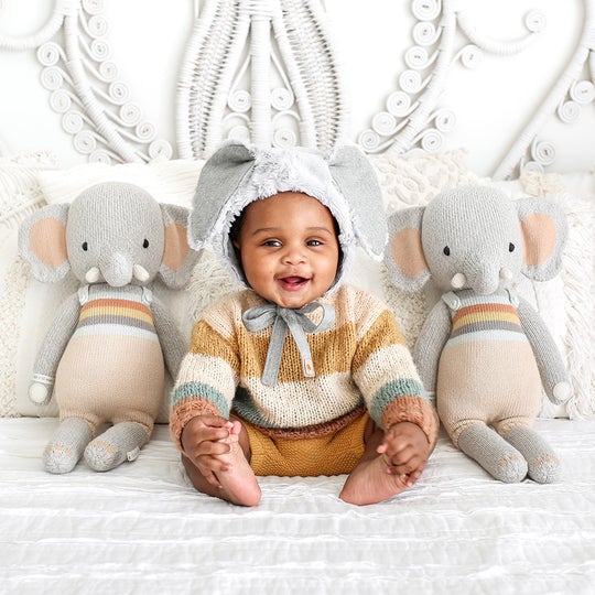 Close up of a baby boy sitting up, wearing an elephant ear bonnet, with 2 Evan The Elephants by Cuddle and Kind, one on either side of him.