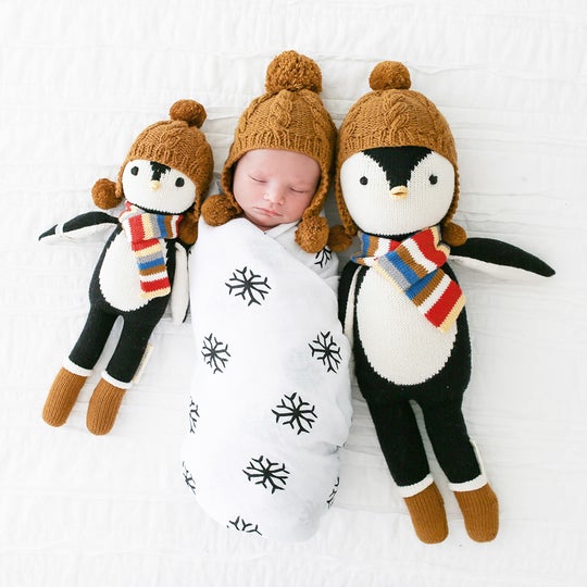 White background with baby sleeping, swaddle in a toque, between both sizes of Everest The Penguin by Cuddle and Kind