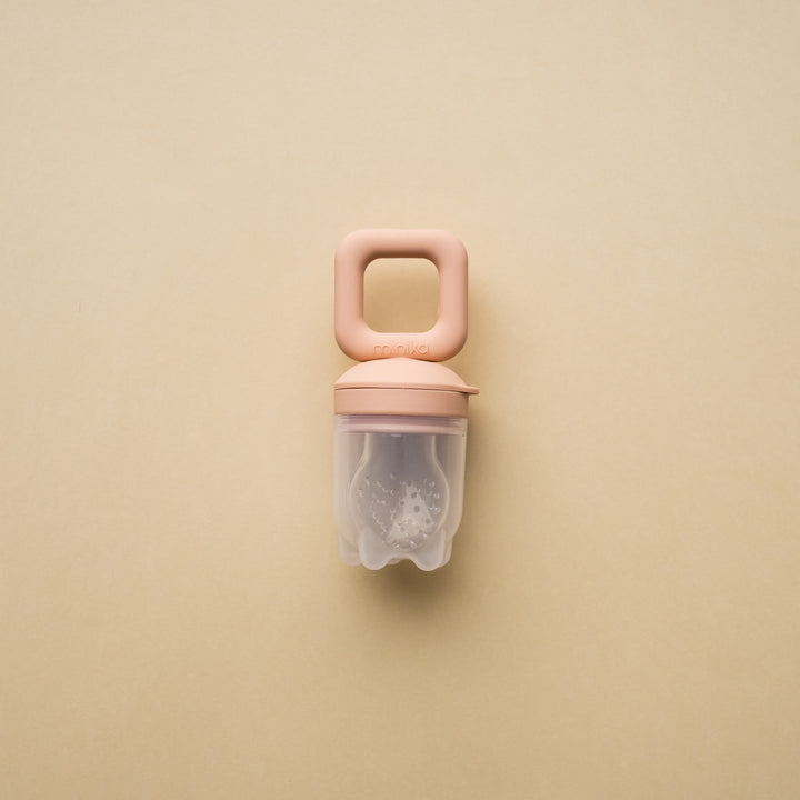 Beige background with a Silicone Feeder Teether in Blush by Minika. Feeder teether has a blush silicone handle, and the feeder part is clear silicone with small holes.