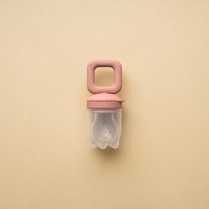 Beige background with a Silicone Feeder Teether in Sorbet by Minika. Feeder teether has a rose speckled silicone handle, and the feeder part is clear silicone with small holes.