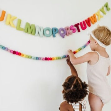 White wall with 2 kids hanging up the Felt Alphabet Garland by The Whimsical Woolies.
