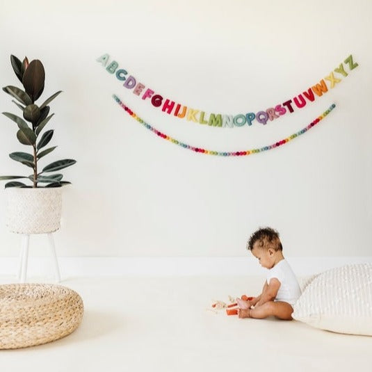 White wall with a a plant on the left, and a baby sitting on the floor, and the Felt Alphabet Garland in Whimsy by The Whimsical Woolies hanging on the wall.