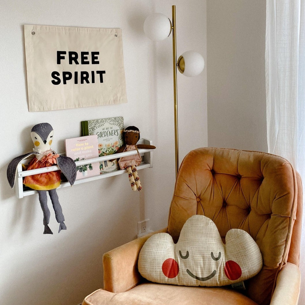 Corner of a nursery, with floating shelf, and a chair, and the Free Spirit Banner by Imani Collective. Banner is linen with the words "FREE SPIRIT" in black.