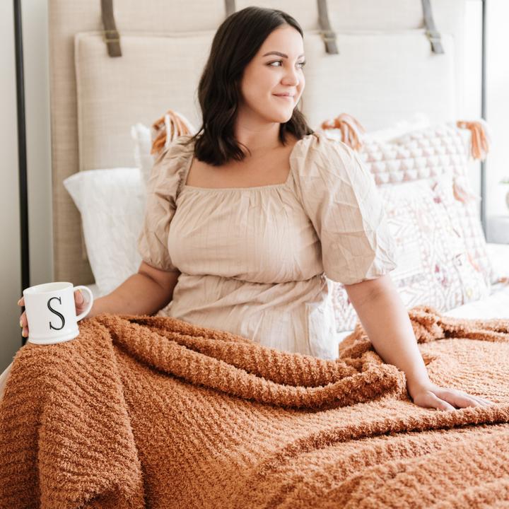 Woman sitting on the edge of her bed with a coffee mug in her hand, and the Ginger Ribbed Bamboni King Size Blanket by Saranoni on her lap. Blanket is a terracotta colour.
