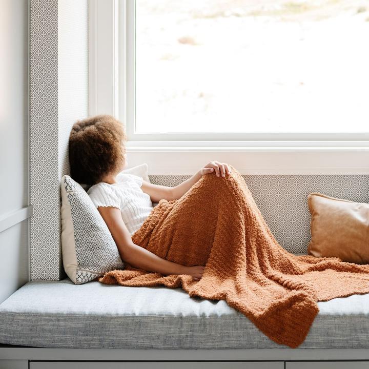 Little girl sitting on a couch beside a window, with the Ginger Ribbed Bamboni Receiving Blanket by Saranoni. Blanket is a terracotta colour.