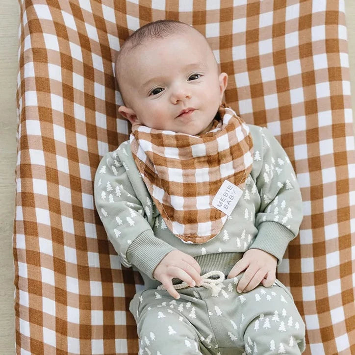 Overhead view of a baby laying down wearing the Gingham Bib by Mebie Baby. Gingham bib is a brown and white gingham.