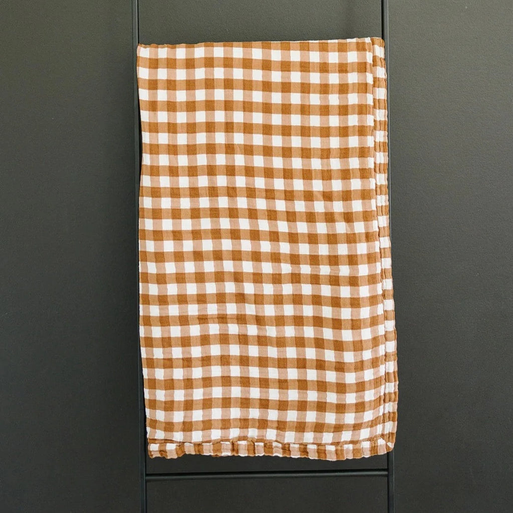 Dark background with a black metal blanket ladder, and the Gingham Muslin Quilt by Mebie Baby hanging off it. Quilt is a brown and white gingham pattern.