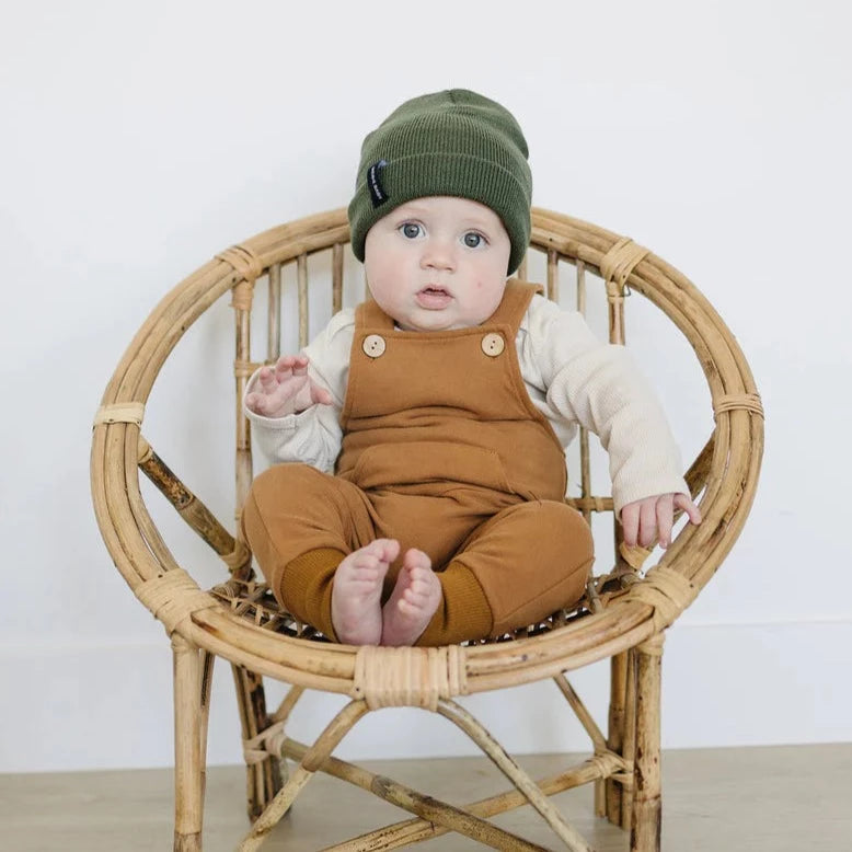 White background with a rattan chair, and a baby sitting on it, wearing a Green beanie by Mebie Baby.
