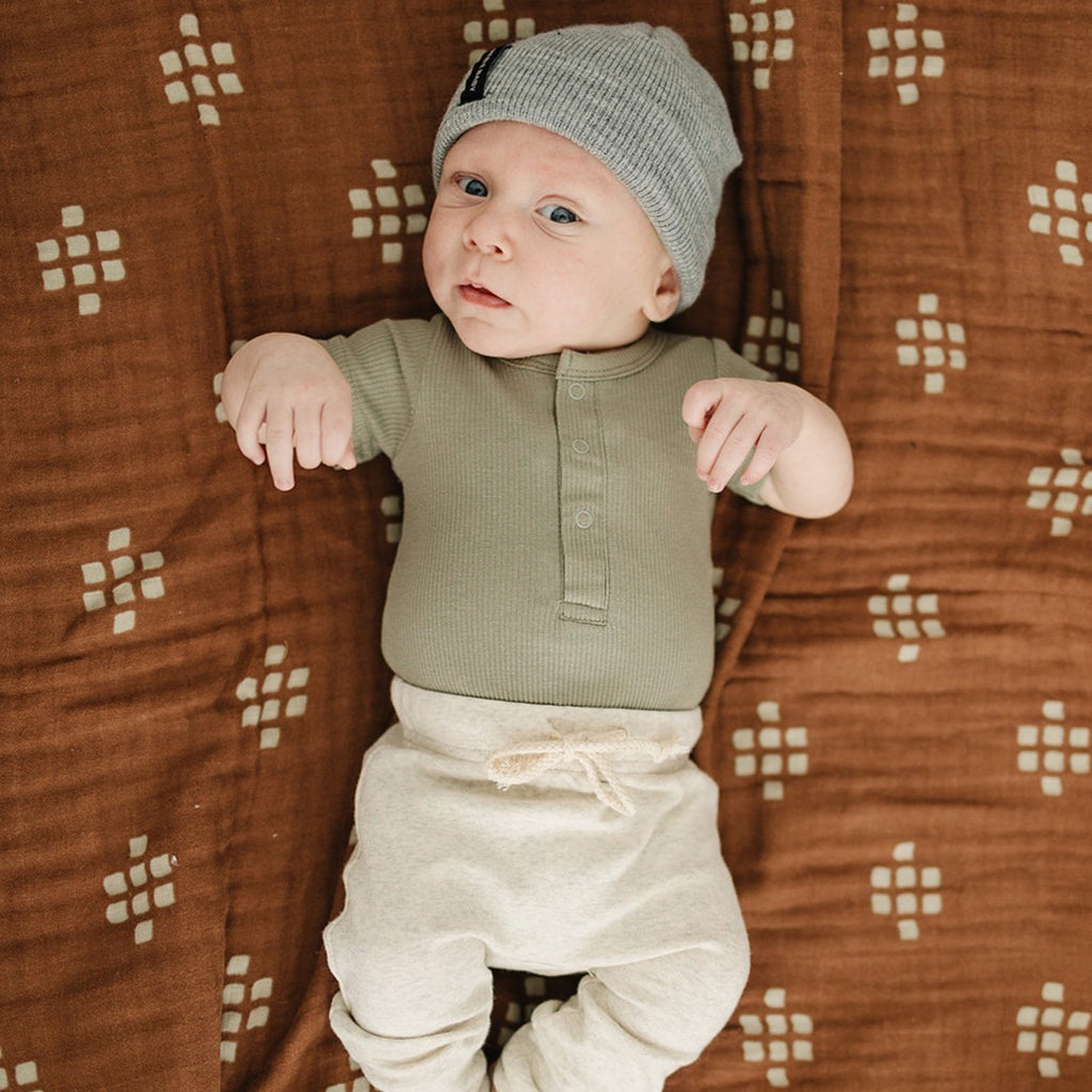 Green Organic Bodysuit by Mebie Baby on a baby wearing pants, Mebie Baby grey hat. Baby is laying on a brown swaddle by Mebie Baby. 
