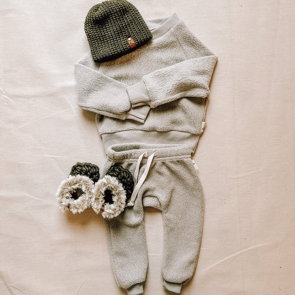 Grey Winter Sky crew and pants by Petit Nordique, paired with olive and fur moccs and an olive beanie by Petit Nordique. Laid on a flat beige surface. 