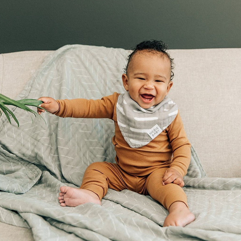 Baby wearing a Grey Dash Bib by Mebie Baby, sitting on a couch with a desert sage swaddle underneath. Baby is holding onto green plant leaf and the baby is wearing a rust coloured outfit, while sitting on a couch in front of a green wall. 