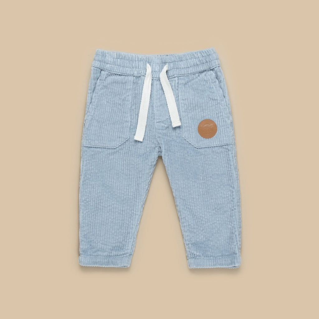 Beige background with Dusty Blue Cord Pant by Hux Baby. Pants are a corded material in a light blue colour, with a white drawstring and a light brown patch on the side.