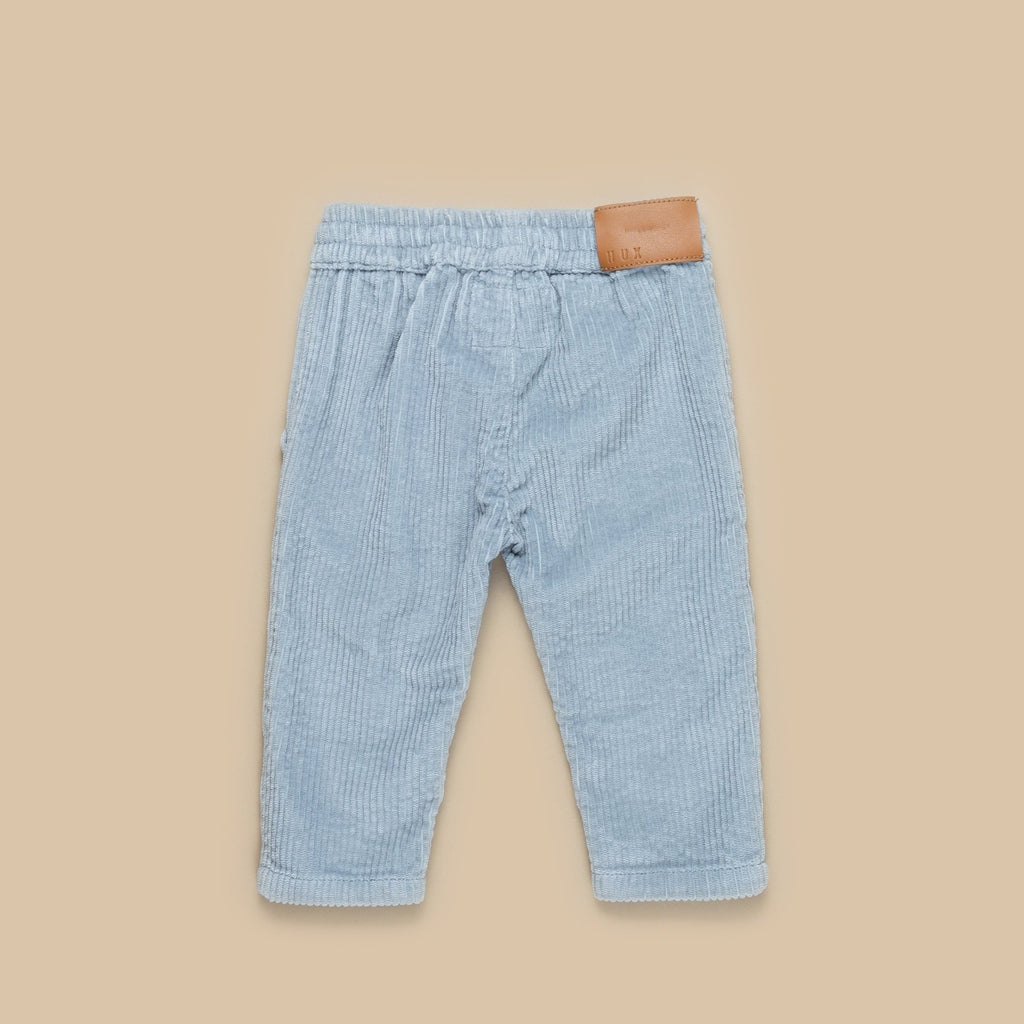 Beige background showing backside of Dusty Blue Cord Pant by Hux Baby. Pants are a corded material in a light blue colour, with a white drawstring and a light brown patch on the side.