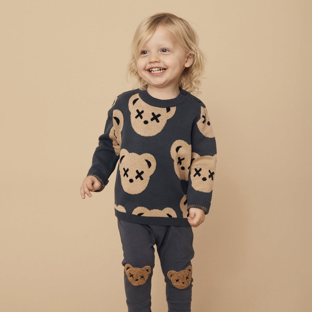 Beige background with boy standing, wearing Huxbear Teddy Knit Jumper in Ink (navy) by Hux Baby. Jumper is navy with light brown bear faces all over, and x's on the eyes.