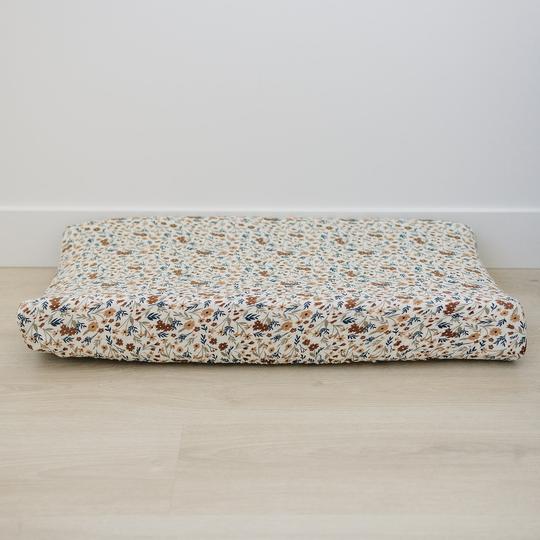 White wall with light wood floor and a change pad with Harvest Floral Changing Pad Cover by Mebie Baby on it. Changing pad cover is a cream/white with flowers in rust, blue, and beige all over, and fits snug to change pad.