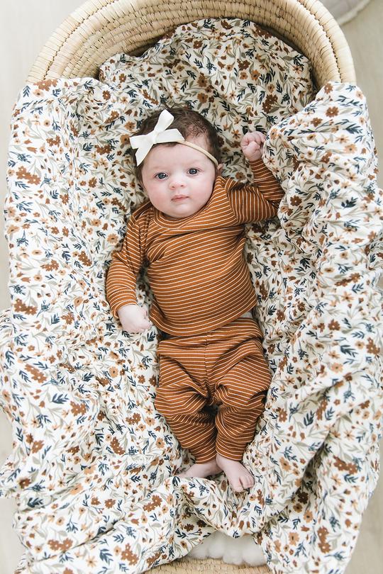 Overhead view of baby laying in a bassinet, with a Harvest Floral Muslin Quilt by Mebie Baby underneath her. Quilt is white/cream with a pattern of flowers in rust, blue, and beige.