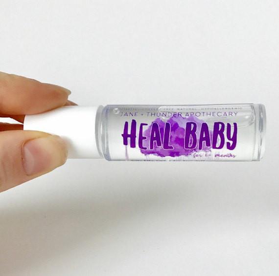 White background with a hand holding Heal Baby - Baby Safe Essential Oil by Jane + Thunder. Writing of "Heal Baby" is in purple font.